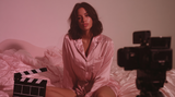A woman in a pink satin pajama top sits on a bed and looks at the camera. In the foreground, a professional video camera is pointed at her, and a movie clapperboard rests on the bed. The scene is lit with a soft pink light.